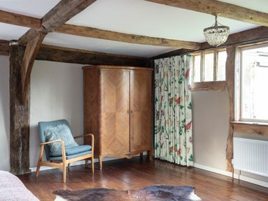 A room with wooden beams a wooden wardrobe a blue armchair with a pillow floral curtains and a window at Brook Farm in St Nicholas near Halesworth