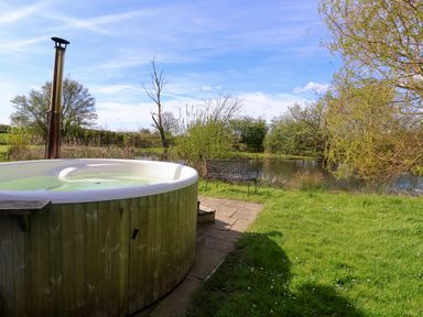 An outdoor hot tub next to a pond with a metal bench on grass at Brook Farm in St Nicholas near Halesworth