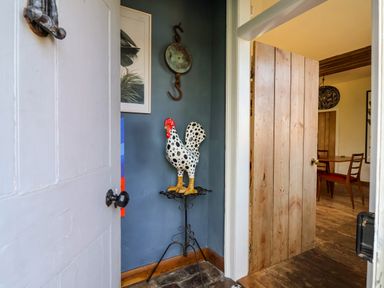 A corner with a decorative rooster statue on a metal stand next to an open wooden door showing a dining area at Brook Farm in St Nicholas near Halesworth