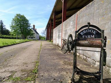 An outdoor farm scene with a weathered farm sign reading Brook Farm next to a concrete path and a large tree near a white house at Brook Farm in St Nicholas near Halesworth
