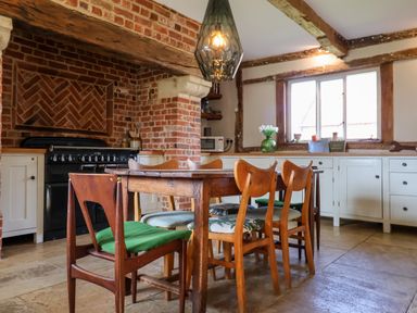 A kitchen with a wooden dining table and chairs near a brick fireplace and white cabinets at Brook Farm in St Nicholas near Halesworth