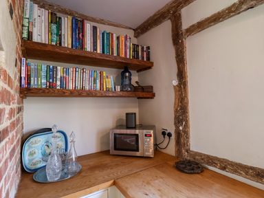 A corner kitchen area with wooden shelves filled with books a microwave on a wooden counter glass bottles on a tray and exposed wooden beams at Brook Farm in St Nicholas near Halesworth
