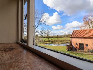 A view through a window showing a wooden windowsill leafless trees a pond a grassy field and a red brick building with a tiled roof at Brook Farm in St Nicholas near Halesworth