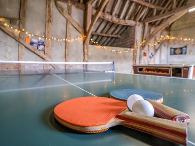 A ping pong table with paddles and balls in a room with wooden beams and fairy lights at Brook Farm in St Nicholas near Halesworth