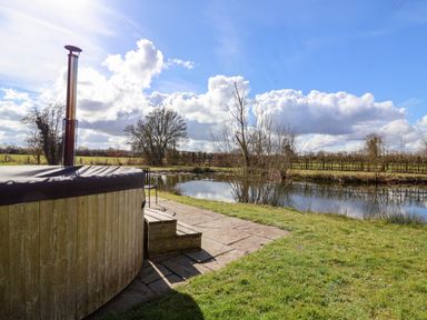 An outdoor wooden hot tub by a pond with grass and trees around at Brook Farm in St Nicholas near Halesworth