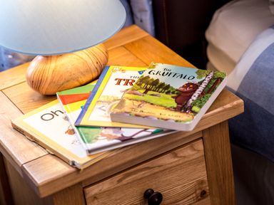 A wooden bedside table with a lamp and a stack of children's books at Mallard in Wheatacre