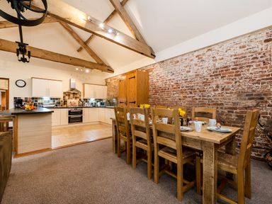 A kitchen and dining area with a wooden table set with dishes and wooden chairs and exposed brick wall at Mallard in Wheatacre
