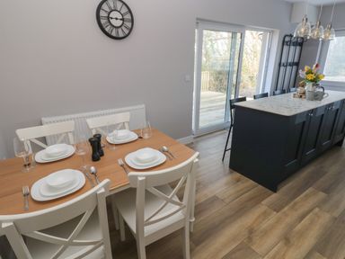 A dining area with white chairs and tableware next to a kitchen island with stools and glass pendant lights at Wychwood in Hook near Haverfordwest