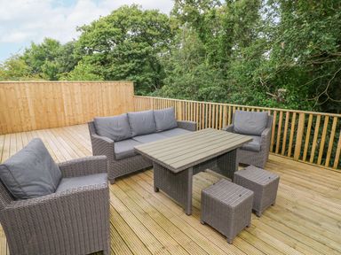 An outdoor wooden deck with wicker furniture including a sofa armchairs stools and a table surrounded by trees at Wychwood in Hook near Haverfordwest