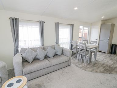 A living room with a grey sofa and cushions next to a dining table with four chairs and windows with curtains at Long Acres Lodge in Evesham