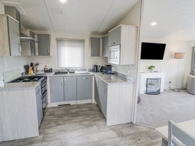 A compact kitchen with gray cabinets and appliances next to a living room with a wall-mounted TV at Long Acres Lodge in Evesham