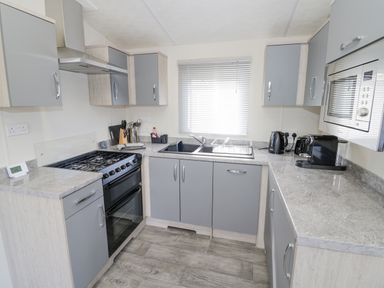 A kitchen with grey cabinets a gas stove a microwave kettle toaster and coffee machine at Long Acres Lodge in Evesham
