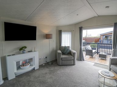 A living room with a wall-mounted TV a white electric fireplace an armchair and a floor lamp next to sliding glass doors leading to an outdoor seating area at Long Acres Lodge in Evesham