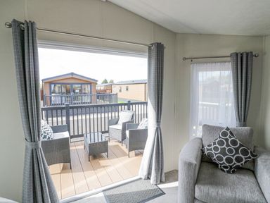 A living area with gray armchair and patterned cushions next to a sliding door leading to a deck with outdoor chairs and table at Long Acres Lodge in Evesham