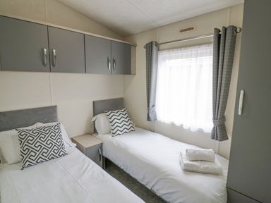 A small bedroom with two single beds with patterned pillows a window with curtains and overhead cabinets at Long Acres Lodge in Evesham