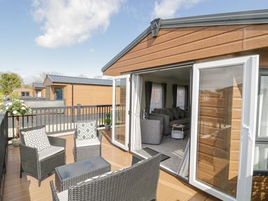 An outdoor deck with wicker furniture and cushions next to an open door leading to a living room at Long Acres Lodge in Evesham