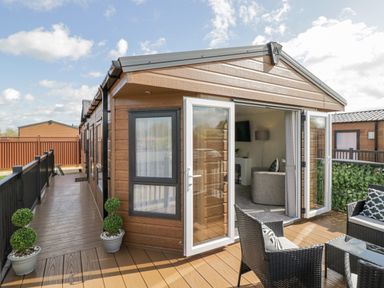 A wooden lodge with open glass doors leading to a deck with outdoor seating and potted plants at Long Acres Lodge in Evesham