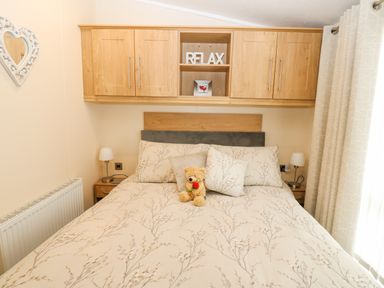 A bedroom with a bed featuring floral bedding a teddy bear bedside lamps wooden overhead cabinets and curtains at Plot 32 in High Hesket near Armathwaite