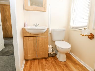 A bathroom with a white toilet a wooden cabinet with a sink a mirror cabinet toilet paper basket and window blinds at Plot 32 High Hesket near Armathwaite