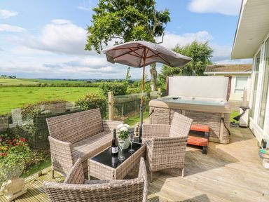 A wooden deck with outdoor wicker furniture a glass table an umbrella a hot tub and a green field in the background at Plot 32 High Hesket near Armathwaite
