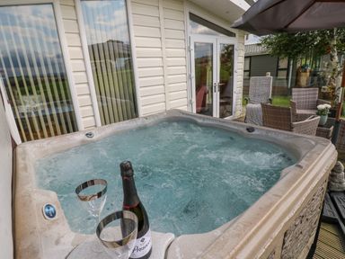An outdoor hot tub with a bottle of prosecco and two glasses near a patio with chairs and tables at Plot 32 High Hesket near Armathwaite