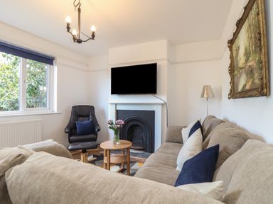 A living room with a beige sofa and blue cushions a black armchair a small round wooden table with flowers a wall-mounted TV and a fireplace at The Heights in Truro