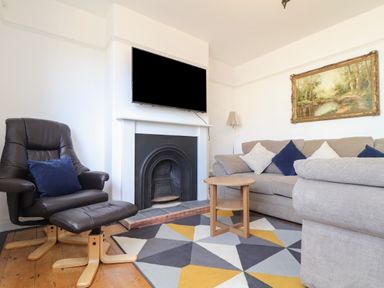 A living room with a beige sofa with cushions a black chair with footrest a fireplace with a TV mounted above a small wooden table and a geometric patterned rug at The Heights in Truro