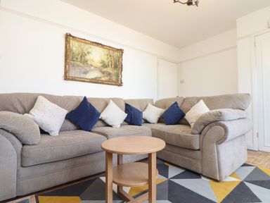 A living room with a beige sectional sofa with white and blue cushions a round wooden coffee table a geometric patterned rug and a framed landscape painting on the wall at The Heights in Truro