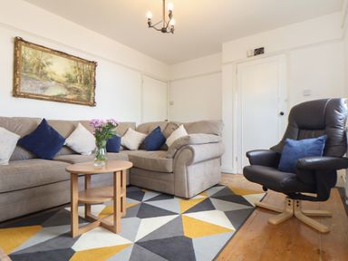 A living room with a beige sectional sofa with cushions, a wooden coffee table with flowers, a black armchair with a cushion, and a geometric patterned rug at The Heights in Truro