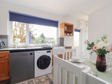 A kitchen with a window above the sink and appliances including dishwasher and washing machine and a dining table with flowers at The Heights in Truro
