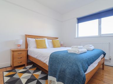 A bedroom with a wooden bed frame a blue blanket two yellow pillows a wooden bedside table with a lamp and a geometric rug at The Heights in Truro