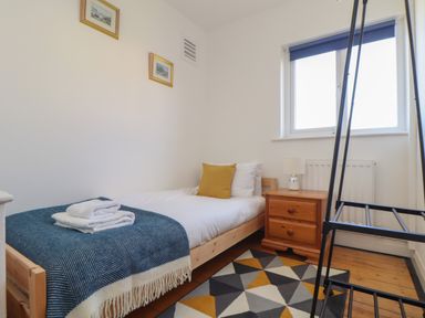 A bedroom with a wooden single bed with white bedding and a yellow cushion and a folded blanket on it beside a wooden bedside table with a lamp and a window with blue blind at The Heights in Truro