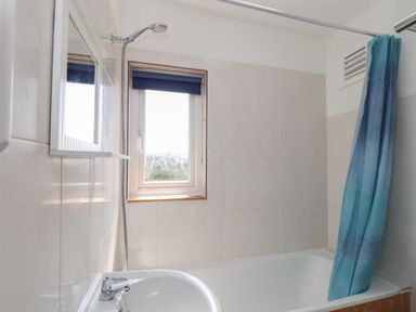 A bathroom with a bathtub shower combination a blue shower curtain a window with a blind and a sink at The Heights in Truro