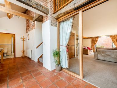 A hallway with terracotta floor tiles and a wooden coat rack next to a bathroom and stairs leading to an upper floor with a living room with sofas and curtains visible through a sliding glass door at Coot in Wheatacre