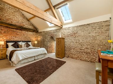 A bedroom with exposed brick walls wooden beams two skylights a bed with bedside tables a brown rug a wooden chest of drawers and a wooden desk at Coot Wheatacre