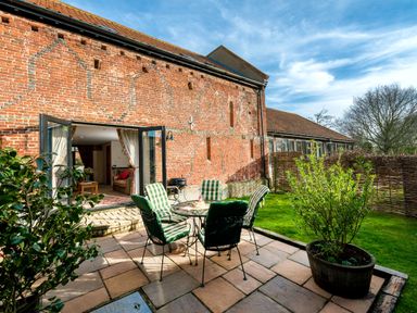 An outdoor patio with a table and chairs next to a brick building with open doors leading into a living room at Coot in Wheatacre