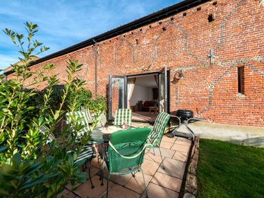 An outdoor patio with green cushioned chairs around a table next to a brick wall with open doors leading to a living room at Coot in Wheatacre