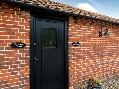 A black door set in a red brick wall with signs reading Fitness Barn and Soft Play Barn and a wall light above a plant pot at Coot in Wheatacre