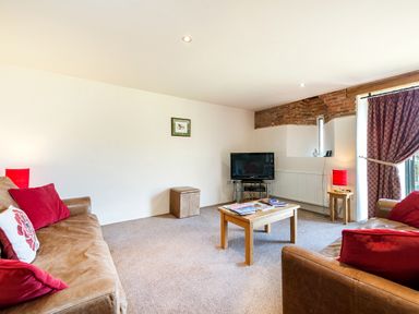 A living room with two brown sofas with red cushions a wooden coffee table a TV on a stand and a side table with a red lamp at Widgeon in Wheatacre