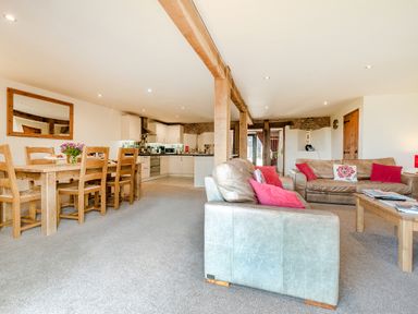 An open plan living room and kitchen with wooden dining table and chairs a leather sofa and armchair with red cushions and exposed wooden beams at Widgeon in Wheatacre