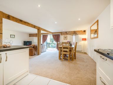 An open plan kitchen with white cabinets leading to a dining area with a wooden table and chairs next to a living area with brown sofas and a TV at Widgeon in Wheatacre