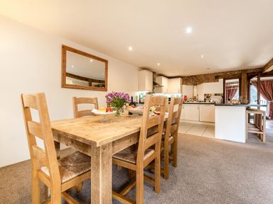 A wooden dining table with six chairs and flowers on the table in a kitchen with white cabinets and an open doorway at Widgeon in Wheatacre