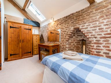 A bedroom with a wooden wardrobe chest of drawers desk chair and a bed with blue checkered bedding against a brick wall at Widgeon in Wheatacre
