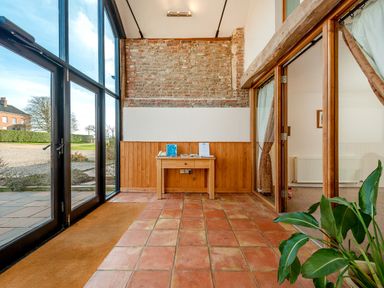 A hallway with terracotta tiled floor a wooden table against a brick wall glass doors on one side and a plant near an open wooden framed glass door at Teal in Wheatacre