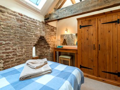 A bedroom with a bed covered by a blue and white checkered blanket towels on the bed a wooden wardrobe a dressing table with a stool and a mirror exposed brick wall with a skylight at Teal in Wheatacre