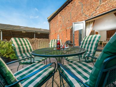 A patio with green and white checkered cushioned chairs around a round metal table with a bottle and glasses near a brick wall at Teal in Wheatacre