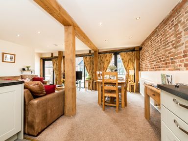 An open living and dining area with wooden beams a brown leather sofa a wooden dining table and chairs and a brick wall at Woodcock in Wheatacre