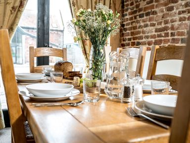 A wooden dining table set with white plates bowls glasses cutlery and a vase of flowers in a room with brick walls at Woodcock in Wheatacre