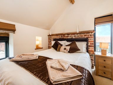 A bedroom with a bed with brown pillows and towels on a brown runner and two windows with lamps Wooden chest of drawers at Woodcock Wheatacre