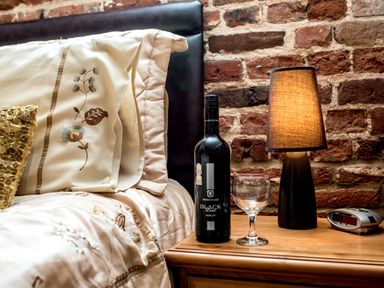 A bed with embroidered pillows next to a wooden nightstand with a bottle of wine a wine glass a lamp and an alarm clock against a brick wall at Woodcock in Wheatacre